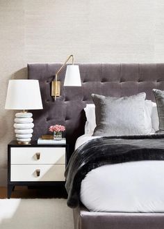 Bedroom features an extra wide gray tufted headboard lit by a brass headboard sconce with gray and white bedding and a black and white nightstand illuminated by a stacked marble lamp.