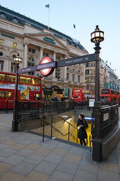 people are walking down the street in front of red double - decker buses at dusk