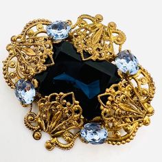 Beautiful Vintage WEST GERMANY Sapphire Blue Glass & Rhinestone Brooch. Ornate gilt filigree metal set with large center sapphire blue glass and accented with light blue rhinestones. Not signed but is known West Germany. In excellent vintage condition with minimal age appropriate wear. Measures 2 inches long by 1 7/8 inches wide. Roll over clasp secure. Formal Blue Gemstone Brooch, Formal Blue Gemstone Brooches, Antique Blue Jewelry With Brooch, Blue Art Deco Formal Brooch, Art Deco Blue Brooches As Gifts, Art Deco Blue Brooches For Gifts, Elegant Blue Gemstone Brooch, Elegant Blue Gemstone Brooches, Elegant Blue Jeweled Brooches