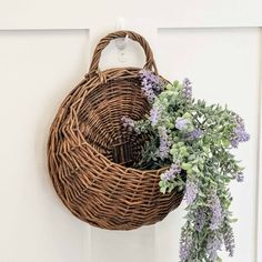 a wicker basket hanging on the front door with purple flowers and greenery in it