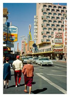 three people walking down the street in front of some buildings and neon signs with cars driving by