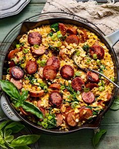 a skillet filled with pasta and sausage on top of a wooden table next to basil leaves