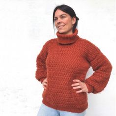 a woman standing in front of a white wall wearing an orange sweater and jeans with her hands on her hips