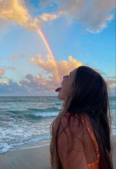 a woman standing on top of a beach next to the ocean with a rainbow in the sky