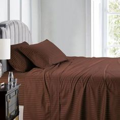 a bed with brown sheets and pillows next to a night stand in front of a window