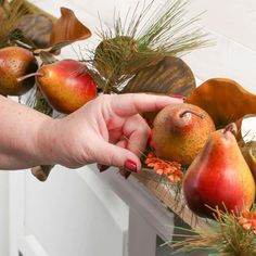 a hand reaching for some fruit on a table with pine cones and flowers in the background