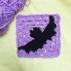 a purple crocheted square with a black bird on it next to a ball of yarn