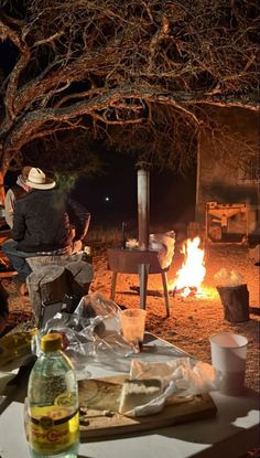 a man sitting at a table in front of a campfire with food on it