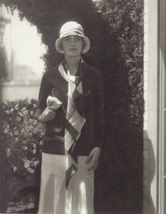 an old photo of a woman wearing a hat and scarf standing in front of a bush