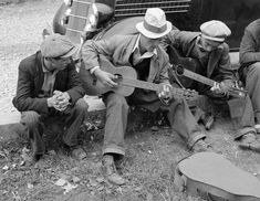 four men sitting on the ground with guitars and hats around their necks, one man is holding a guitar