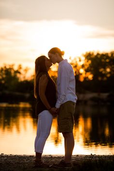 a man and woman standing next to each other near the water at sunset with trees in the background