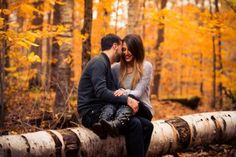 a man and woman sitting on a log in the woods with autumn leaves around them