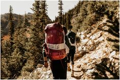 two people hiking up a mountain with backpacks on their back and trees in the background