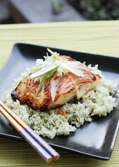 a black plate topped with chicken and rice next to chopsticks on a table