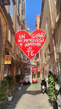 a red heart shaped sign hanging from the side of a building next to parked motor scooters