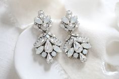 a pair of white crystal earrings sitting on top of a white cloth next to a flower