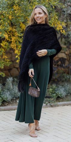 a woman in green pants and a black shawl smiles while holding a brown purse