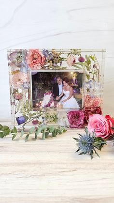 a wedding photo frame with flowers and greenery on the table, next to it is a bouquet of roses