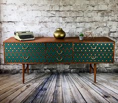 an old fashioned desk with a green and gold patterned design on the front, against a white brick wall