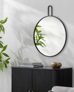 a round mirror hanging on the side of a wall next to a black cabinet and plant