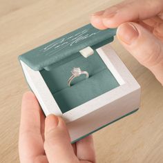 a person holding an engagement ring in a box on top of a wooden table with writing on it