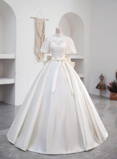 Laced with the nostalgia of a bygone era, this wedding dress boasts a high-neck lace bodice that radiates elegance and poise. Its delicate sleeves brush the edges of the shoulders, a soft prelude to the voluminous satin skirt that cascades into a regal train. The simplicity of the silhouette is its grandeur, tied together by a satin sash at the waist, making this gown a timeless choice for a bride's unforgettable day. Unique Ball Gowns, Silver Decorations, Satin Long Prom Dress, Lace Top White, Formal Wedding Dress, डिजाइनर कपड़े, Top In Pizzo, Satin Formal Dress, Short Sleeve Wedding Dress