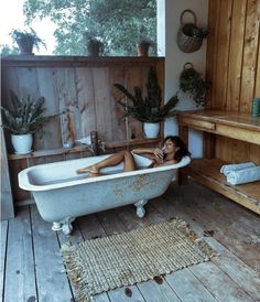 a woman laying down in a bathtub next to potted plants and wooden shelves