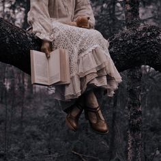 a woman is sitting on a tree branch with an open book in her lap and holding a teddy bear