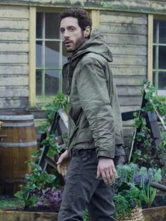 a man standing in front of a garden filled with flowers and vegetables, wearing a green jacket