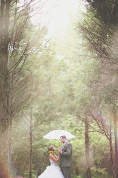 a bride and groom standing under an umbrella in the rain with trees behind them at their wedding