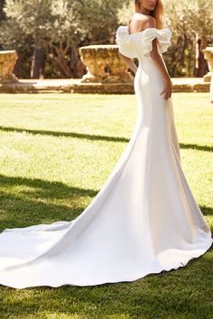 a woman in a white wedding dress standing on the grass with her arms behind her back