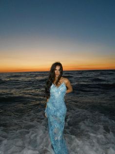 a woman in a blue dress standing on the beach at sunset with her arms behind her back