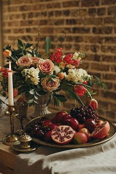 flowers and fruit on a table in front of a brick wall with candlesticks