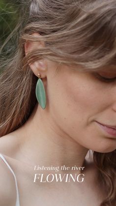 a woman with long hair wearing large green earrings and a white tank top is looking down at her ear