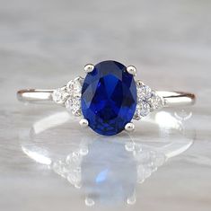 ♥ Beautifully handcrafted Blue Sapphire ring that features 1.5-carat classic oval cut Sapphire gemstone with brilliant-cut Diamond accents (5A grade CZ) in solid Sterling Silver. ♥ This ring makes the perfect engagement or anniversary gift for her that will be treasured forever! ♥ Blue Sapphire is traditionally associated with love and purity. It's said to bring prosperity and attract gifts of all kinds. Metal: Solid 925 Sterling Silver Gemstone: Blue Sapphire Side Stones: Diamond simulants (5A Blue Rings With Side Stones And Round Cut, Blue Round Cut Rings With Side Stones, Silver Oval Rings With Side Stones, Oval Silver Rings With Side Stones, Blue Oval Birthstone Ring For Anniversary, Oval Blue Birthstone Ring For Anniversary, Blue Jewelry With Side Stones In Round Cut, Blue Round Cut Jewelry With Side Stones, Classic Blue Cubic Zirconia Birthstone Ring