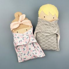 two dolls are sitting next to each other on a gray background, one is wearing a blanket and the other has a pink bow