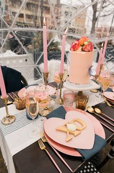 a table set up with pink and gold plates, candles, and cake in the shape of a heart