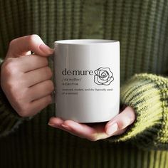 a woman holding a white coffee mug in her hands with the words demure on it