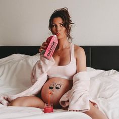 a pregnant woman sitting on a bed drinking from a pink bottle and holding a straw