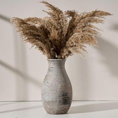 a vase filled with dried grass sitting on top of a white table next to a wall