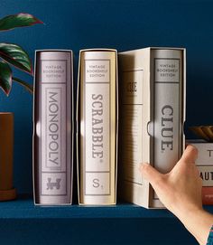 a hand is pointing at three books on a shelf next to a potted plant