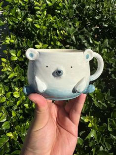 a hand holding up a ceramic mug with a bear on it's side in front of some bushes