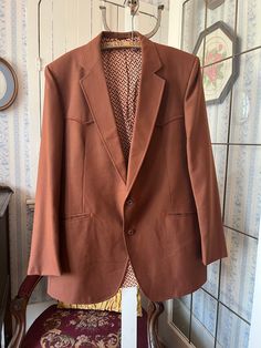 This jacket from Mister Jack's Men's Wear Ltd. is made from a wool blend in rust brown. It has the original buttons in the front and on the cuffs, two front pockets and two inside pockets, and it's fully lined with brown, beige and orange print lining. It has western styling on the front and back yokes and pockets. The measurements, taken with the coat lying flat, are: shoulder to shoulder, 18 inches; armpit to armpit, 23 inches; sleeves, 27 inches; length, 36  inches; bottom edge, 34 inches. There is a small light spot on the right lapel (see photo), but otherwise the coat is in very good condition. Brown Single Button Blazer, Brown Tweed Jacket With Single Button, Brown Single Button Long Sleeve Tweed Jacket, Brown Single Button Long Sleeve Suit, Brown Tweed Jacket With Single Button And Lapel Collar, Brown Blazer With Single Button And Lapel Collar, Brown Single Breasted Blazer With Notch Lapel, Brown Fitted Outerwear With Single Button, Fitted Brown Outerwear With Single Button