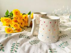 two mugs sitting on top of a bed next to yellow flowers