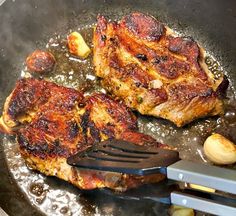 two pieces of meat cooking in a frying pan with spatula and tongs