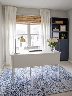 a white desk sitting in front of a window next to a blue and white rug