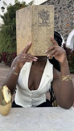 a woman sitting at a table with a book in her hand and wearing gold bracelets