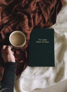 a person holding a coffee cup next to a book on top of a white blanket