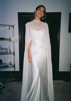 a woman in a white gown and cape standing next to a wall with shelves on it