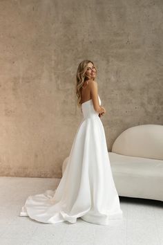 a woman standing in front of a white couch wearing a strapless wedding dress with an open back